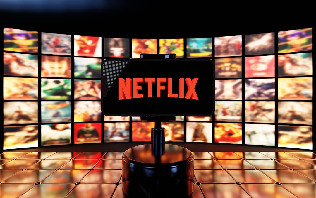 A smartphone displays the Netflix logo in bright red, set against a background of vibrant, colorful movie thumbnails and a glossy tiled surface. The cinematic atmosphere highlights streaming culture and digital entertainment.